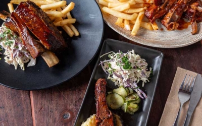 Settlers_Tavern_Food_OBJ886794 A variety of rib dishes on plates with fries, coleslaw, and mashed potatoes, set on a wooden table with drinks.