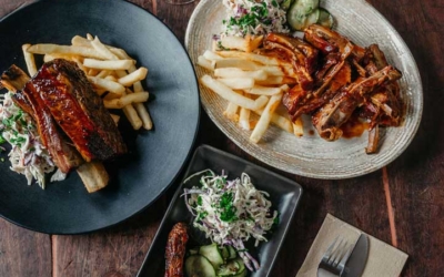 Settlers_Tavern_Food_OBJ886796 Three plates of ribs with fries and coleslaw on a wooden table, accompanied by a glass of white wine, red wine, and a pint of beer.