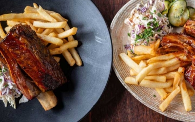 Settlers_Tavern_Food_OBJ886801 Three plates of food on a wooden table: ribs with fries and slaw, ribs with mashed potatoes and slaw, and a pint of beer in a glass.