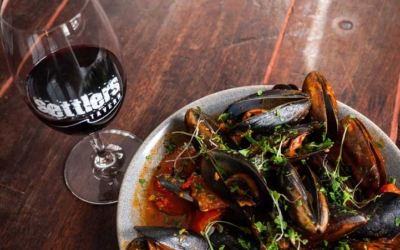 Settlers_Tavern_Food_OBJ886803 A plate of mussels in a red sauce garnished with herbs, next to a glass of red wine on a wooden table.