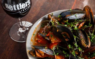 Settlers_Tavern_Food_OBJ886806 A plate of mussels in tomato sauce garnished with herbs next to a glass of red wine on a wooden table.