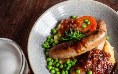 Settlers_Tavern_Food_OBJ886808 A plate of sausages with mashed potatoes, peas, and a tomato slice. Topped with gravy and rosemary. A glass of beer is on the wooden table beside it.