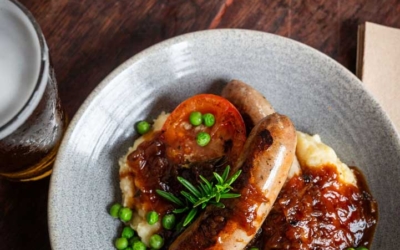 Settlers_Tavern_Food_OBJ886810 A dish of sausages on mashed potatoes with peas and gravy, garnished with a tomato slice, next to a glass of beer on a wooden table.
