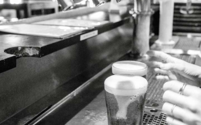 Settlers_Tavern_Food_OBJ886855 Black and white image of beer taps with a pint glass being filled with beer on a bar counter. Hands are visible adjusting the glass.