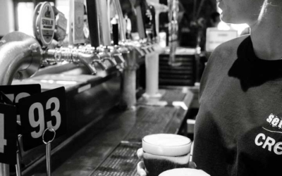 Settlers_Tavern_Food_OBJ886857 A person holding two pints of beer stands near a bar with multiple beer taps in a dimly lit setting.