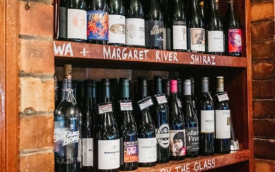 Settlers_Tavern_Food_OBJ886865 Wooden shelves displaying various wine bottles with labels and rows of empty wine glasses beneath, set against a brick wall.