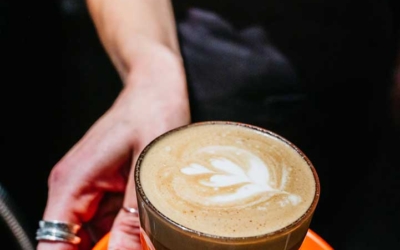 Settlers_Tavern_Food_OBJ886871 A hand holds a glass of latte with latte art on an orange saucer, accompanied by a spoon and a piece of brown sugar.