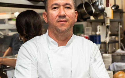 Settlers_Tavern_Food_OBJ886905 A chef stands confidently with arms crossed in a busy kitchen, wearing a white chef's coat and a wristwatch.