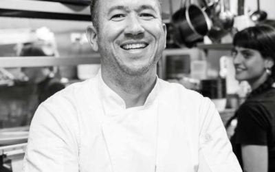Settlers_Tavern_Food_OBJ886909 A smiling chef in a white uniform stands in a professional kitchen with arms crossed. Cooking utensils hang in the background.