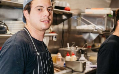 Settlers_Tavern_Food_OBJ886919 A man in a kitchen wearing a striped apron and a backward cap stands next to another person. Shelves with plates and kitchen supplies are in the background.