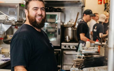 Settlers_Tavern_Food_OBJ886921 A man with a beard smiles while standing in a busy restaurant kitchen, holding a utensil near a fryer. Other kitchen staff work in the background.