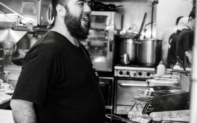 Settlers_Tavern_Food_OBJ886922 Chef standing in a commercial kitchen, wearing a black T-shirt, next to various cooking equipment.