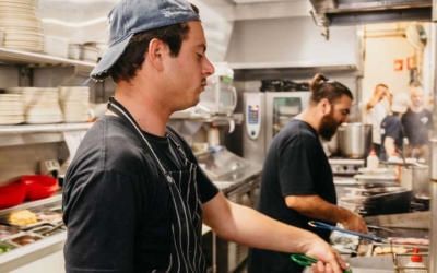 Settlers_Tavern_Food_OBJ886923 Two chefs work in a busy restaurant kitchen, using deep fryers. One wears a blue cap and striped apron while the other is in black.
