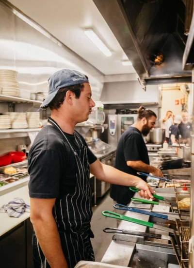 Settlers_Tavern_Food_OBJ886923 Two chefs work in a busy restaurant kitchen, using deep fryers. One wears a blue cap and striped apron while the other is in black.