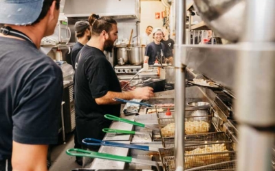 Settlers_Tavern_Food_OBJ886929 Chefs work in a busy kitchen, preparing food at a row of fryers. Some are wearing black shirts and caps.