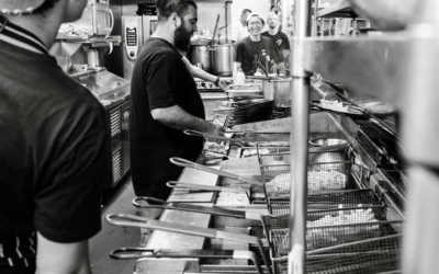 Settlers_Tavern_Food_OBJ886930 Black and white photo of a restaurant kitchen with three staff members cooking and preparing food at the fryers.