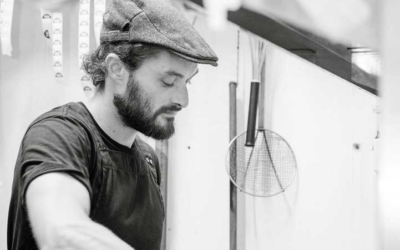 Settlers_Tavern_Food_OBJ886939 A man in a flat cap works in a kitchen, holding metal containers. Cooking utensils hang on the wall behind him.