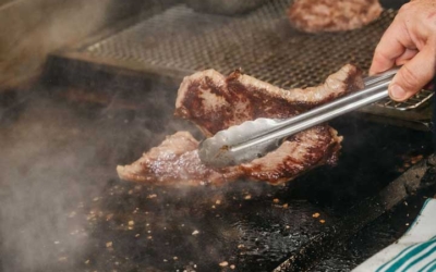 Settlers_Tavern_Food_OBJ886951 A hand uses tongs to flip a steak on a hot grill, surrounded by steam. Another patty is seen cooking nearby. A blue-striped towel is in the foreground.