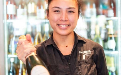 Settlers_Tavern_Food_OBJ887088 A person smiling and holding a bottle of champagne in front of a refrigerated display of various bottles.
