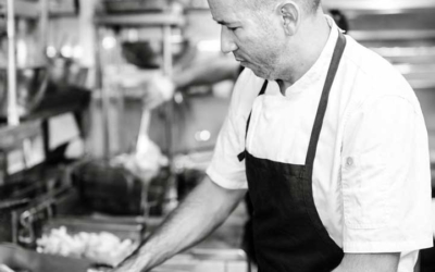 Settlers_Tavern_Food_OBJ887091 Chef in a kitchen wearing a black apron, focused on cooking at the stove.