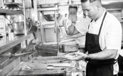 Settlers_Tavern_Food_OBJ887094 Chef in a professional kitchen sprinkling seasoning over a pan, surrounded by cooking equipment and utensils.