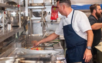 Settlers_Tavern_Food_OBJ887104 A chef wearing a white shirt and blue apron cooks on a busy commercial kitchen stovetop with several pans. Another person in the background works at the counter.