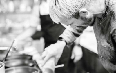 Settlers_Tavern_Food_OBJ887106 A chef with light hair focuses on seasoning a dish in a busy kitchen, alongside another chef working in the background.