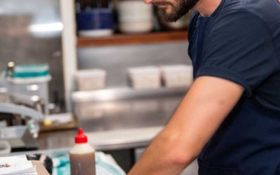 Settlers_Tavern_Food_OBJ887132 A man wearing a cap and apron prepares salad in a stainless steel bowl in a kitchen.