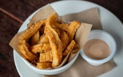 Settlers_Tavern_Food_OBJ887149 A bowl of fried churros on paper in a white dish, accompanied by a small cup of light brown dipping sauce, placed on a wooden table.