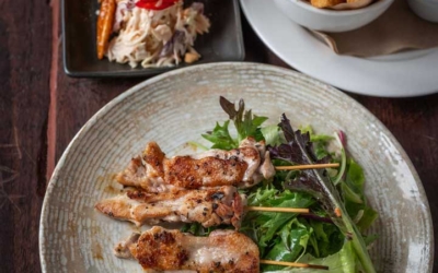 Settlers_Tavern_Food_OBJ887159 A plate of grilled chicken with mixed greens in the foreground, a dish of onion rings, and a platter of fried foods in the background on a wooden table.