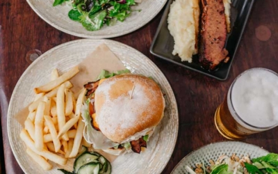 Settlers_Tavern_Food_OBJ887169 An overhead view of a table set with various dishes, including a burger with fries, salad, steak with coleslaw, and a glass of beer.