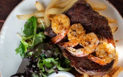 Settlers_Tavern_Food_OBJ887212 A plate with grilled steak topped with shrimp, fries, greens, and a small bowl of sauce.