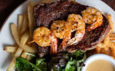 Settlers_Tavern_Food_OBJ887214 A plate with steak topped with shrimp, served with fries, greens, and a small cup of sauce. A knife is placed beside the dish.
