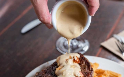 Settlers_Tavern_Food_OBJ887225 A hand pours creamy sauce over steak topped with shrimp, served with fries and a side salad on a wooden table.