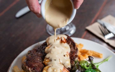 Settlers_Tavern_Food_OBJ887228 A hand pours sauce over a steak topped with shrimp, served with fries and a side salad on a plate.