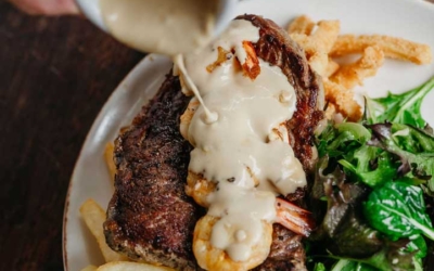 Settlers_Tavern_Food_OBJ887230 A hand pouring sauce over a steak with shrimp, accompanied by fries and a green salad.