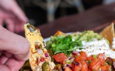 Settlers_Tavern_Food_OBJ887233 A person lifting a nacho covered with melted cheese, jalapeños, diced tomatoes, and onions from a plate. Sour cream and guacamole are also on the dish.