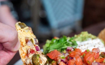 Settlers_Tavern_Food_OBJ887234 A hand holds a nacho chip loaded with cheese, jalapeños, and toppings from a dish of nachos with diced tomatoes, sour cream, and guacamole.