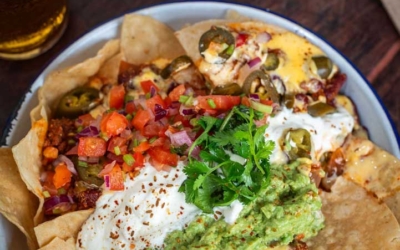 Settlers_Tavern_Food_OBJ887244 A plate of nachos topped with diced tomatoes, jalapeños, guacamole, sour cream, melted cheese, and fresh cilantro, next to a glass of beer on a wooden table.