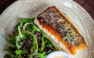 Settlers_Tavern_Food_OBJ887246 A plate with a grilled fish fillet, mixed greens salad, and a small bowl of creamy dressing. A glass of white wine is in the background.