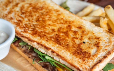 Settlers_Tavern_Food_OBJ887259 Toasted sandwich with greens and meat, served with fries and a side of sauce, next to a tall glass of beer on a wooden table.