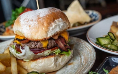 Settlers_Tavern_Food_OBJ887263 A cheeseburger with lettuce, bacon, and pickles on a plate, accompanied by fries and other dishes in the background.