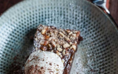 Settlers_Tavern_Food_OBJ887307 A plate with a chocolate brownie topped with a scoop of vanilla ice cream and sprinkled with nuts and cocoa powder. A fork is placed beside the plate on a wooden table.