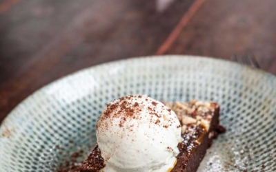 Settlers_Tavern_Food_OBJ887310 A rectangular brownie topped with a scoop of vanilla ice cream and sprinkled with chocolate sits on a patterned ceramic plate on a wooden table.