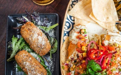 Settlers_Tavern_Food_OBJ887321 A plate of croquettes with greens, a dish with nachos topped with salsa and herbs, and a glass of beer on a wooden table.