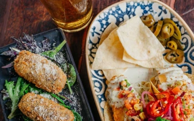 Settlers_Tavern_Food_OBJ887324 A glass of beer is placed next to a plate of three croquettes on greens, and another plate with nachos, jalapeños, and salsa.