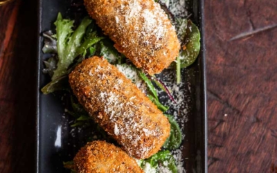 Settlers_Tavern_Food_OBJ887326 Three breaded croquettes topped with grated cheese, served on a bed of mixed greens on a black rectangular plate.