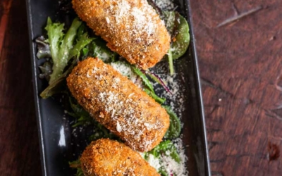Settlers_Tavern_Food_OBJ887330 Three breaded croquettes on a bed of mixed greens, served on a rectangular black plate, sprinkled with cheese, on a wooden table.