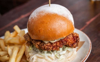 Settlers_Tavern_Food_OBJ887340 A crispy fried chicken sandwich with lettuce on a bun, served with fries and pickles on a wooden table. A glass of beer is in the background.