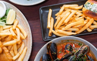 Settlers_Tavern_Food_OBJ887365 A table with plates of sausages with peas, spaghetti, fries, fish and chips, mussels in sauce, and a glass of beer.